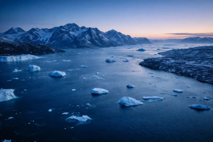 Greenland’s Arctic coastline at blue hour with drifting icebergs in a wide icy fjord beneath snow-covered mountains.